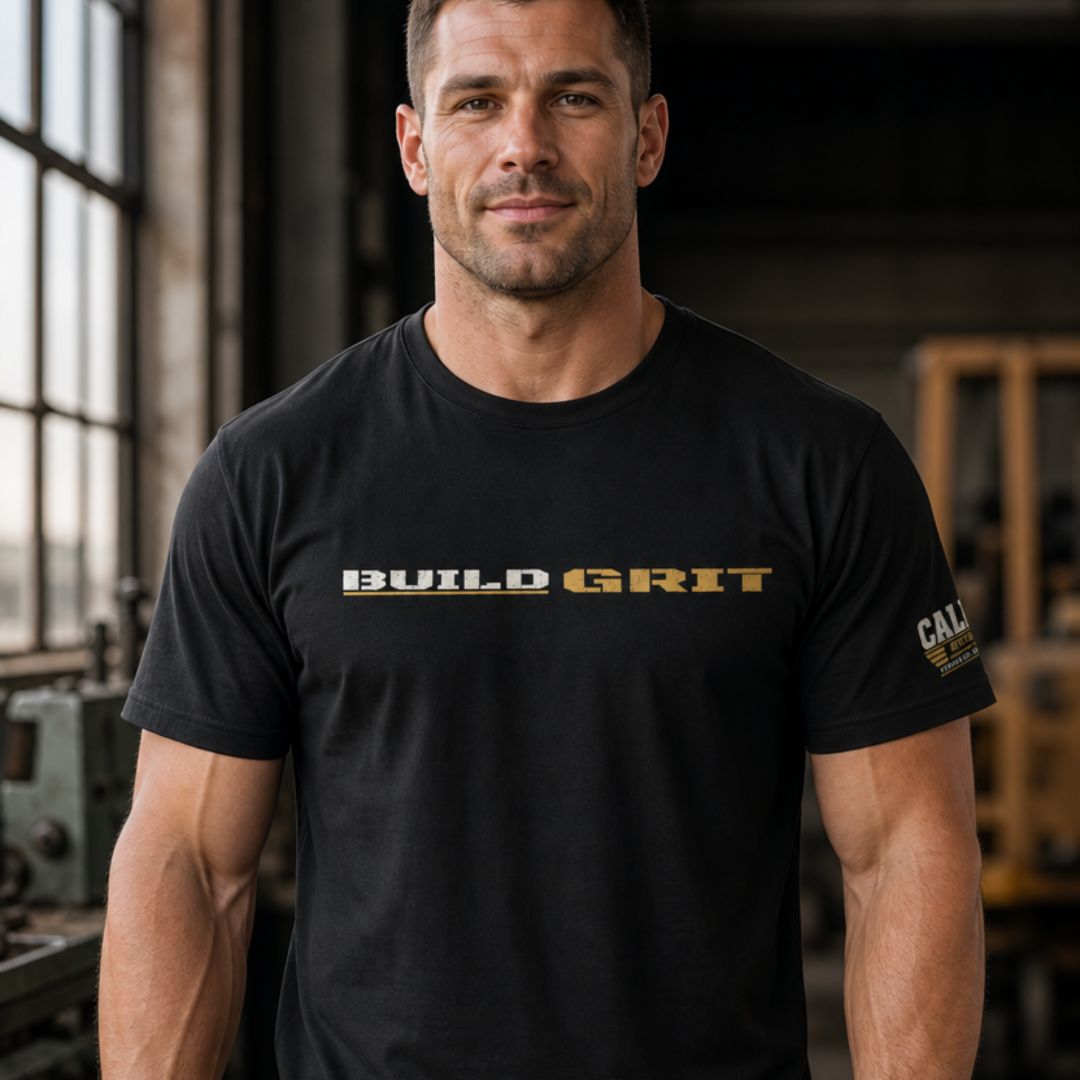 Man wearing the BUILD GRIT Premium T-Shirt in Graphite Black featuring distressed front artwork and a Caliber Motivation Co. sleeve patch.