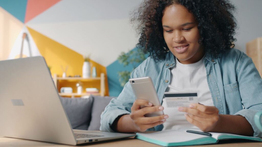 Teen using a laptop, bank card, and smartphone to learn banking, bills, and credit