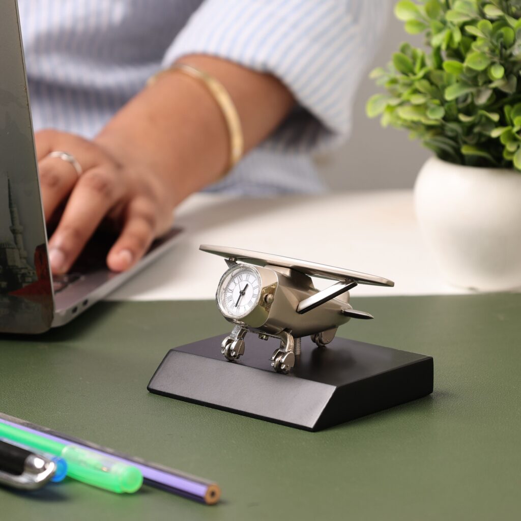 the-now-habit-productivity-book-clock A silver airplane-shaped desk clock resting on a black base on a green desk mat, with a person typing on a laptop blurred in the background.